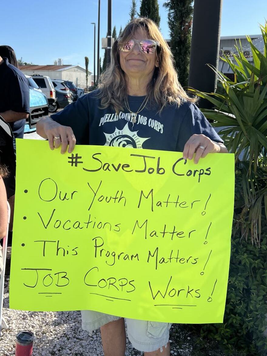An older woman with long brown hair holds a bright yellow sign with handwriting. It says #SaveJobCorps. Our Youth Matter! Vocations Matter! This proram matters! Job Corps Works!