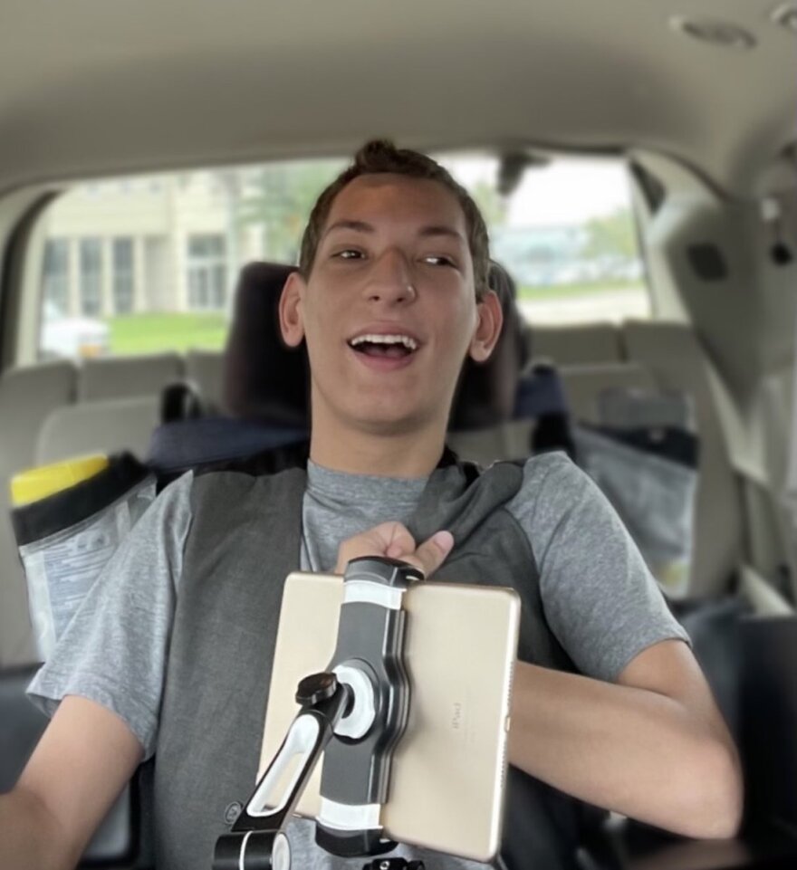 A young man wearing a grey shirt is smiling. He's in a vehicle. 