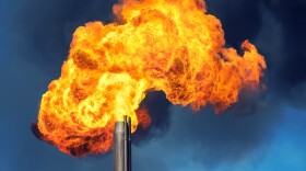 A flare of on-fire gases explodes from a pipe against a blue sky.