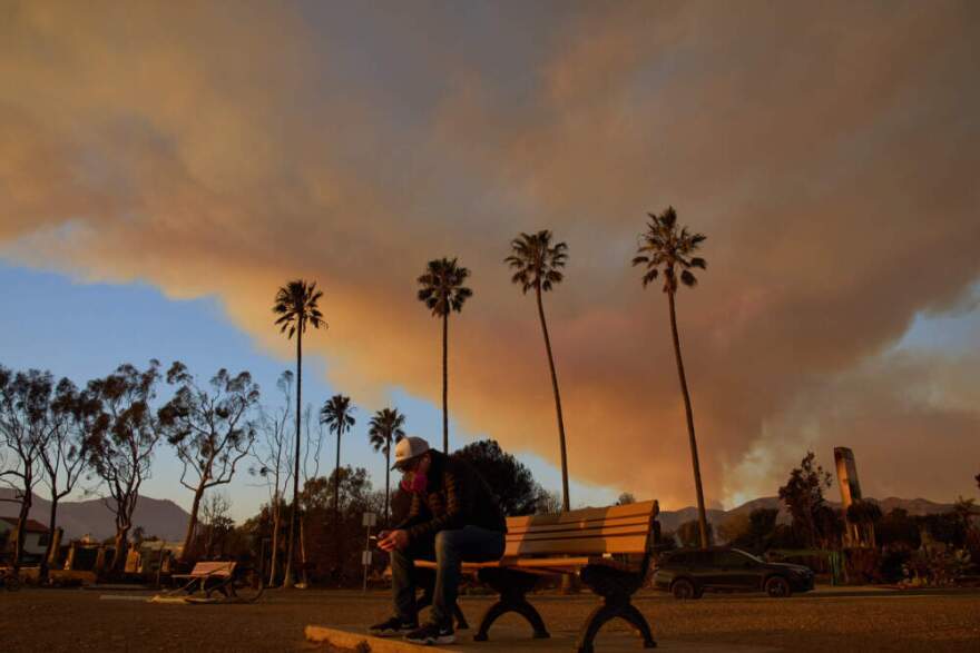 A person sits on a bench as a plume of smoke rises from the Palisades Fire, Friday, Jan. 10, 2025, in Pacific Palisades, California. (John Locher/AP)