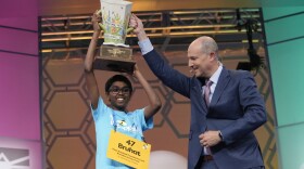 Boy with glasses and large yellow nametag holds up a trophy with a man in a suit helping him