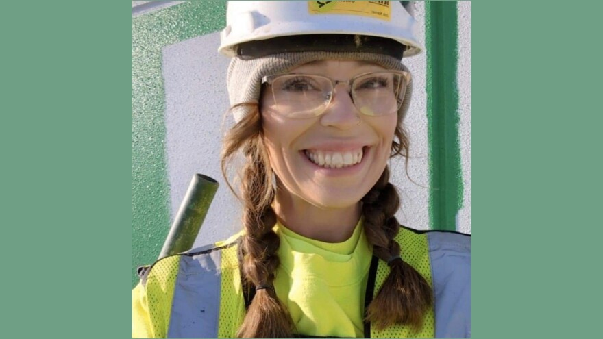 A woman wearing glasses, a white hard hat, and a yellow safety vest smiles at the camera. She has brown hair in braided pigtails and stands in front of a painted wall with green and white sections.