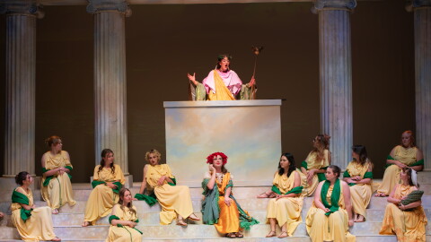 On a stage decorated as a Greek amphitheater, one woman in a Greek costume speaks from a podium to several other women who sit on the steps below. 