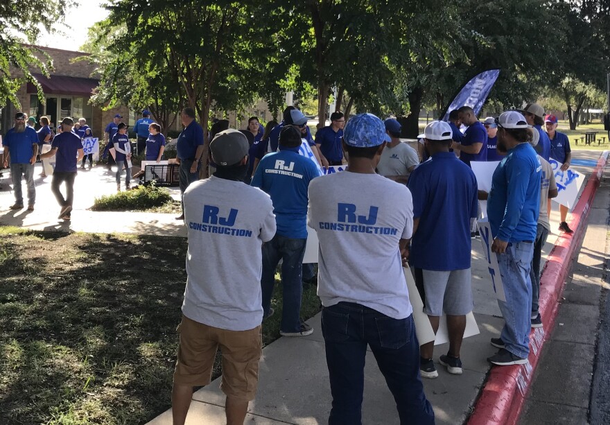 Two men in baseball caps wear white shirts that say "RJ Construction" in blue lettering. In front of them, a sea of friends and family of Robert Jordan wearing blue RJ Construction shirts fill the plaza outside Arlington Independent School District's administrative building, which is surrounded by trees. In the far background, people hold signs that say "Pay RJ."