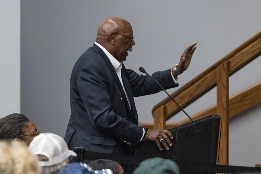 A photo of Rich Stoglin, a Black man with a white mustache wearing a dark suit, gestures as he speaks at a podium.