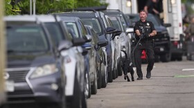 An officer walks a black police K9 along cars parked on the side of a road. In the background, you can see more law enforcement.
