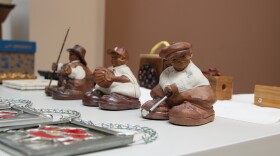 Three clay sculptures of fat men holding different sports equipment sit on a table. In front of them are stained glass red flowers. Behind them are wooden blocks.