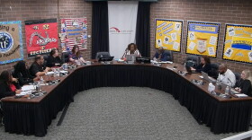 A group of people sit around a large U-shaped table in a meeting room with bulletin boards on the walls and “Peoria Public Schools” on a banner behind the center person.