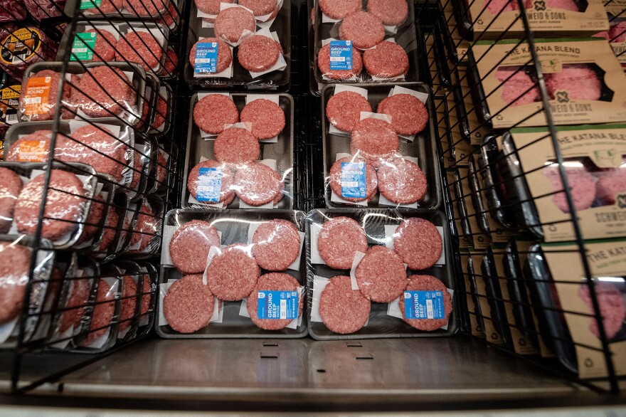 Packaged raw hamburger patties in plastic trays are stacked on shelves in a refrigerated grocery store display.