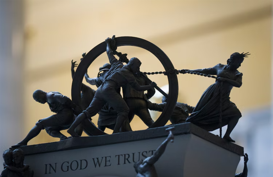 A close up of a mini model of people on top of a platform that reads "In God We Trust." The figures on top of the platform are pulling and pushing a hoop.