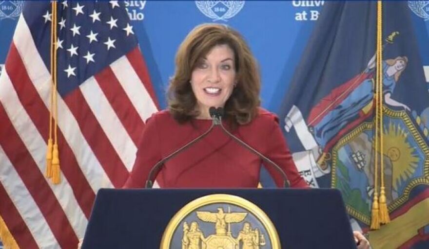 Gov. Kathy Hochul, wearing a red sweater, stands at a navy podium with the gold state seal on it and the U.S. and state flags behind her.