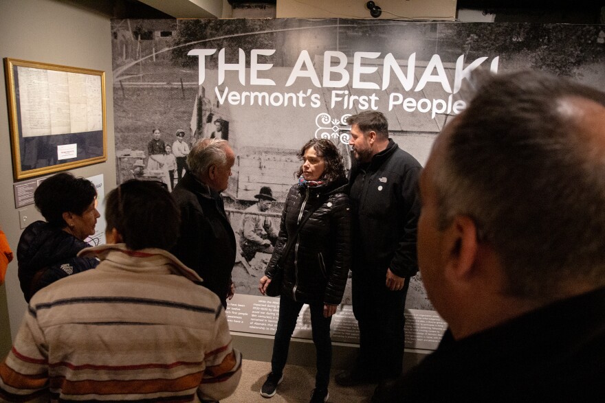 A photo of people crowding into an exhibit space with an enlarged title on the wall behind the people, reading: The Abenaki: Vermont's First People. 