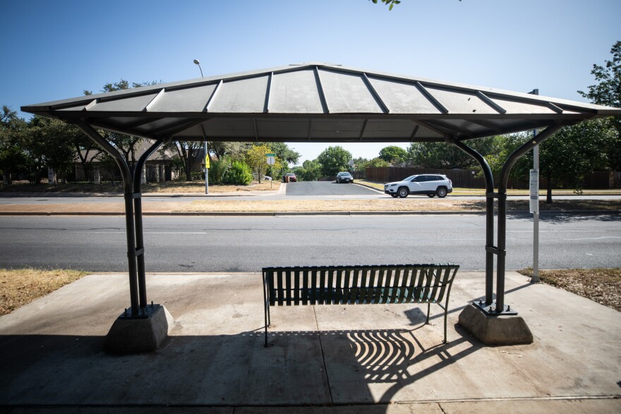 An older CapMetro bus shelter. A six-foot wide bench rests under the partial shade of the canopy. A white SUV is driving by in the background.