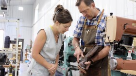A man in a buttoned-down shirt and apron holds up a cowboy boot being stitched together to show a woman in a denim jumper.