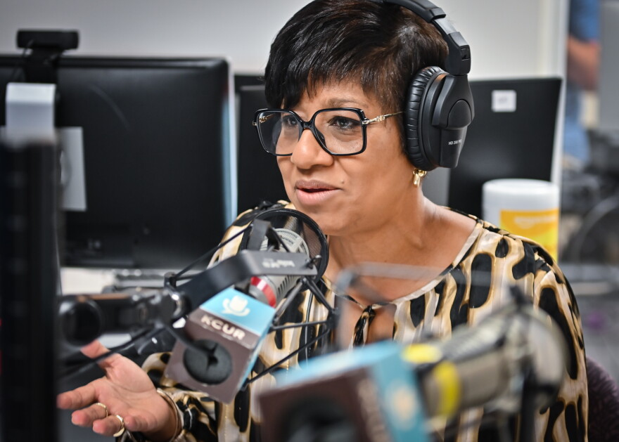 A woman sits inside a radio studio wearing a headset and talking while gesturing with her right hand.