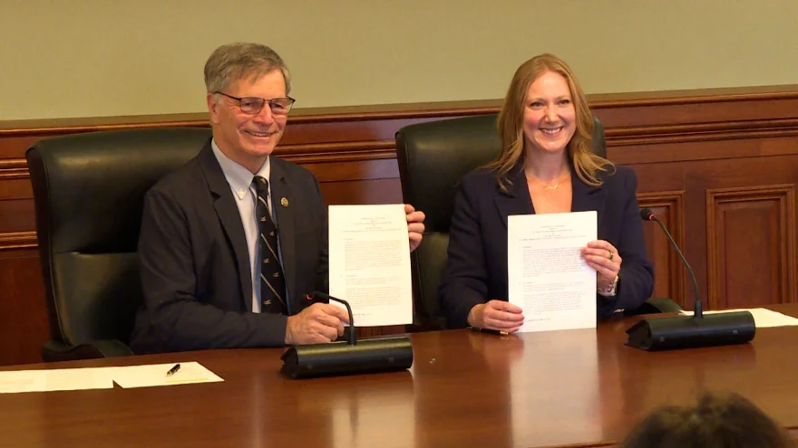 A grey haired man with glasses and a suit holds up a piece of paper. Next to him a blond haired woman holds up a piece of paper. 