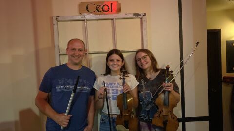 A teenager and an adult are holding fiddles while smiling. A second adult is holding a woodwind instrument while smiling.
