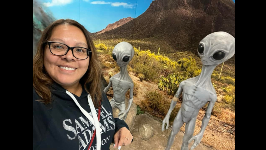 Lady wearing a black hoodie in a museum. She is standing in front of two alien sculptures, posing and taking a selfie. 