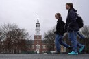 A man wearing blue crocs and a woman walk from the right to the left. In the background, a brick campus building.