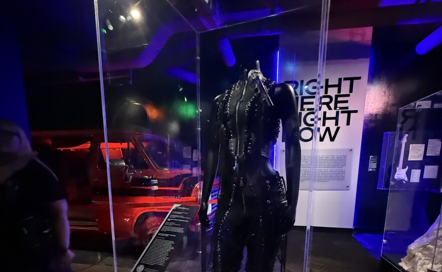 A vertical glass museum case displays a black leather halter bodysuit with silver medal studs. Behind, an orange car is partially visible in the dark lighting.