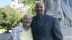 Older woman poses with a man in front of a statue and several trees with a courthouse in the background