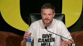 A man answers questions behind a microphone at a desk.