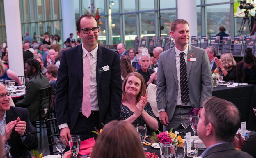 Saint Louis Public Radio Politics Correspondent Jason Rosenbaum and Saint Louis on the Air Executive Producer Alex Heuer stand up at their table at the 2026 Hear the Future gala while the other attendees clap.