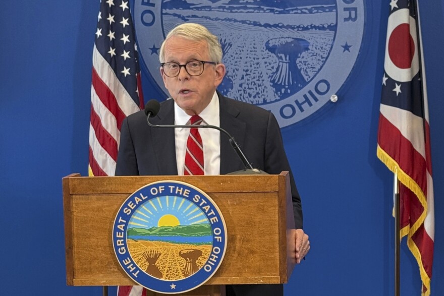 A photo shows a man with glasses standing at a podium. The wood-grained podium has a sign that reads: “The Great Seal of the State of Ohio.”  Behind him is an American flag and a flag of the state of Ohio.