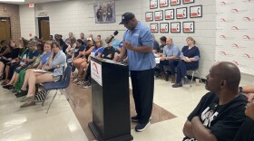 Carl Cannon wears a blue shirt and speaks into a podium surrounded by people in three rows of chairs. 