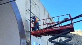 A person stands on a massive hydraulic lift as they work on some electrical wires on the side of a very tall tunnel.