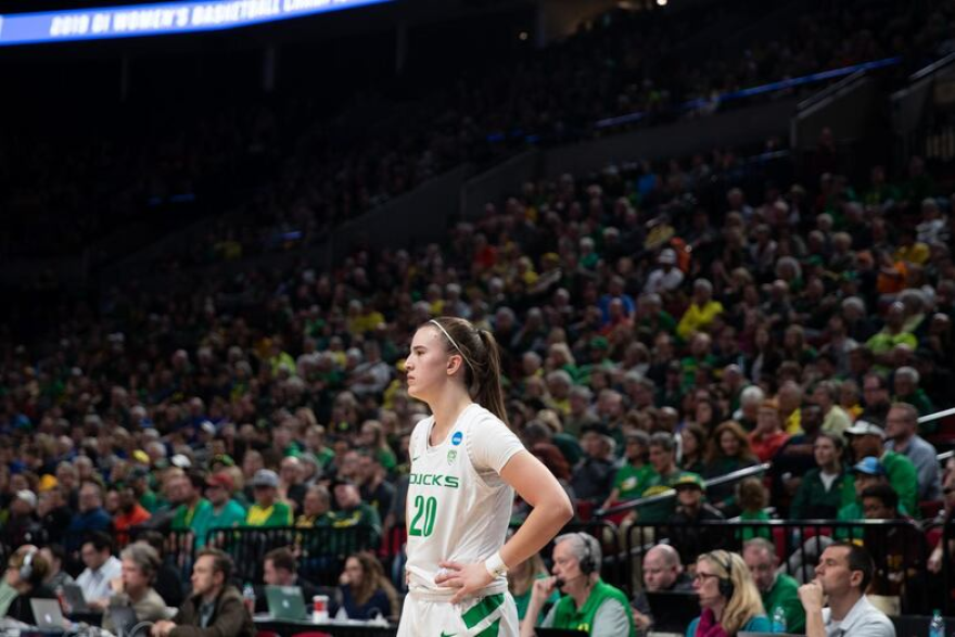 Basketball player Sabrina Ionescu in an Oregon Ducks uniform with a seated crowd behind her.