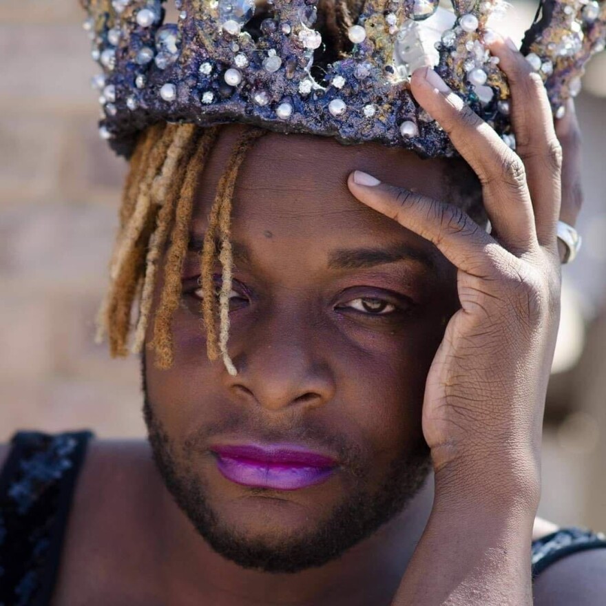 A Black man looks directly at the camera with tired eyes. He's wearing violet lipstick and is wearing his bleached hair in dreadlocks. Atop his head is a glittering fabric crown.