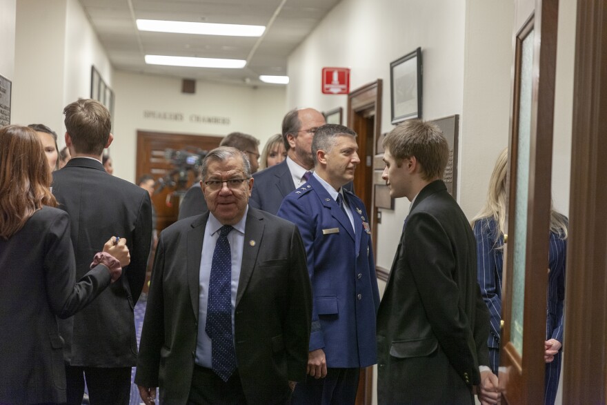 A white man in a suit and blue tie walks throug a crowded corridor