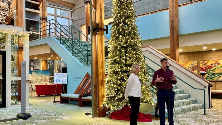 A man and a woman stand in front of a lit holiday tree. The man speaks into a microphone. 
