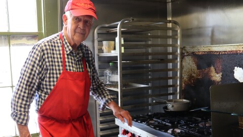 A man wearing a red apron and cap stands beside a stove in a kitchen.