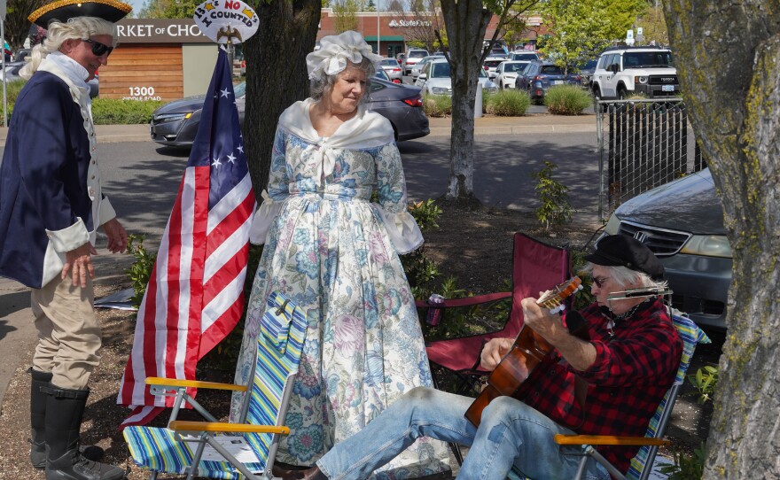 Two people in colonial-era style outfits stand beside an American flag while a seated man plays guitar during a protest along a tree-lined street with parked cars in the background.