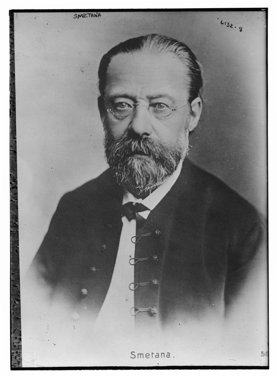 An older man with glasses, a mustache and a beard, dressed in a dark coat with a white shirt, looks toward the camera