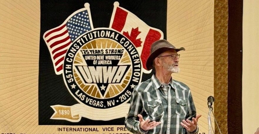 A white older man with a short mustache and beard  wearing a green and white flannel speaks towards a microphone with both of his hands turned up in front of him. In the background is a quilt that says 55th Constitutional Convention Las Vegas, NV 2015 on the wall. There's a backwards American flag to the left and a Canadian flag to the right on the quilt. At the bottom it says 1890 with a quilted ribbon leading towards the right side that the man is standing in front of.