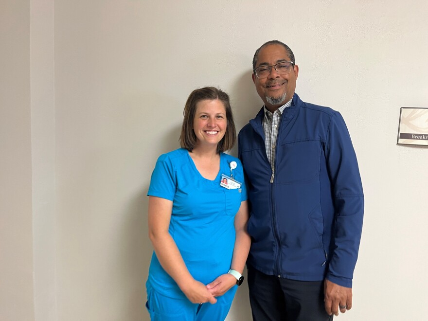 A white woman with medium length brown hair is wearing blue scrubs, is standing beside a Black man with glasses and a long-sleeved blue jacket. 