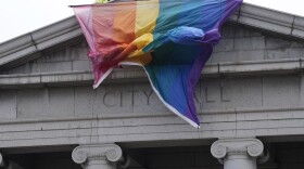 A large rainbow pride flag billows over the top of a gray stone building with columns, engraved at the top with the words "City Hall."