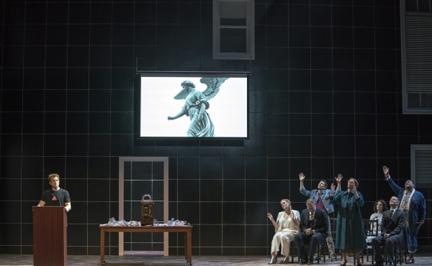 photo of stage production - a man speaks at a podium and a group of people look up at an angel figure