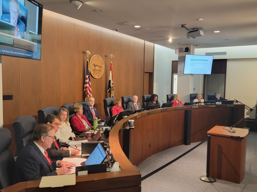 Eleven people sit at a elevated dais. Five are white women, four are white men, and two are Black women. The American and Missouri flags are visible behind them, flanking the seal of St. Louis County.