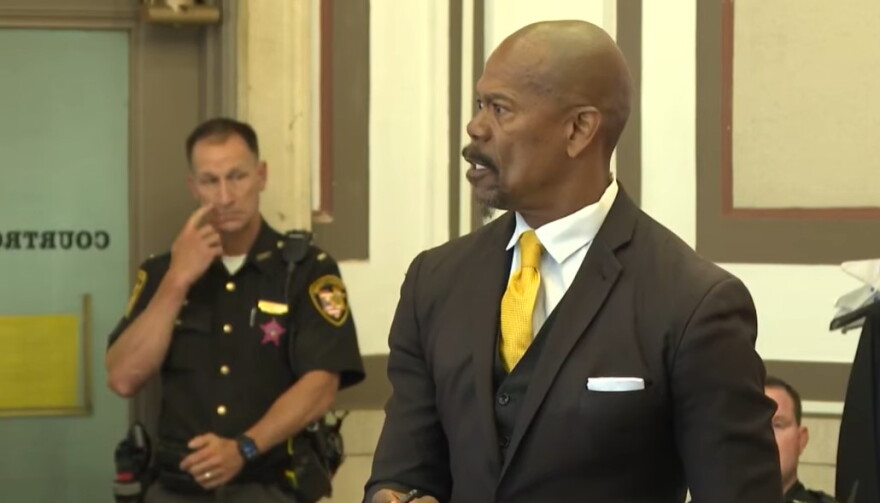 man in suit looks to his right in a courtroom. a sheriff's deputy is seen in the background
