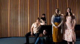 Five people stand or sit around a piano in a music hall building. They are all looking into the camera wit neutral expressions, wearing business casual clothes.