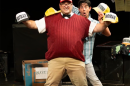 A man in a red sweater vest, a black bowtie, and khakis wears a red trucker hat that says "Monk" and holds two other trucker hats. Behind him, a man in a short sleeved plaid button up holds another trucker hat in one hand and a remote in the other. 