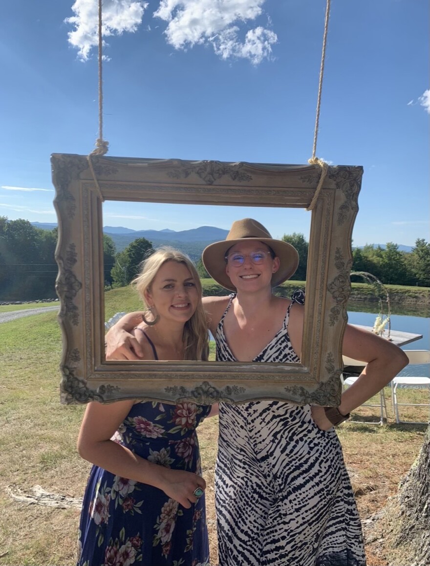 A photo of two light-skinned women standing with their arms around each other, their faces framed by an empty picture frame hanging from ropes. In the background is blue sky and green fields and mountains on a sunny day. The women are wearing strapless dresses, and the woman on the right is wearing a hat and glasses.