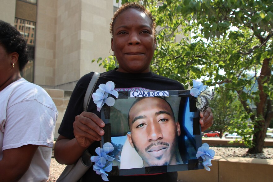 A woman stands holding a picture of a man with the name "Cameron" at the top and blue flowers in the corners.