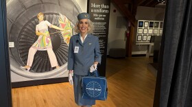A woman wearing a blue vintage outfit with white gloves and a blue Pan Am bag stands at the entrance to a dimly lit room. Behind her is a photo of a woman wearing a pink dress and holding a colorful umbrella while standing in the well of a plane's engine. 