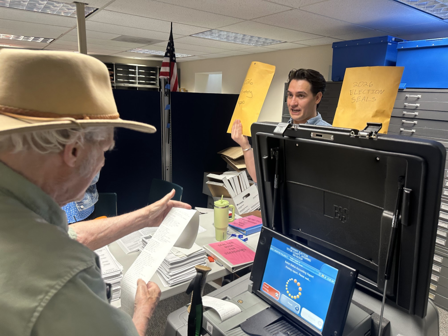 An older man wearing a tan fedora holds a long slip of paper. A younger man standing across a desk from him holds up two large envelopes of election related documents.