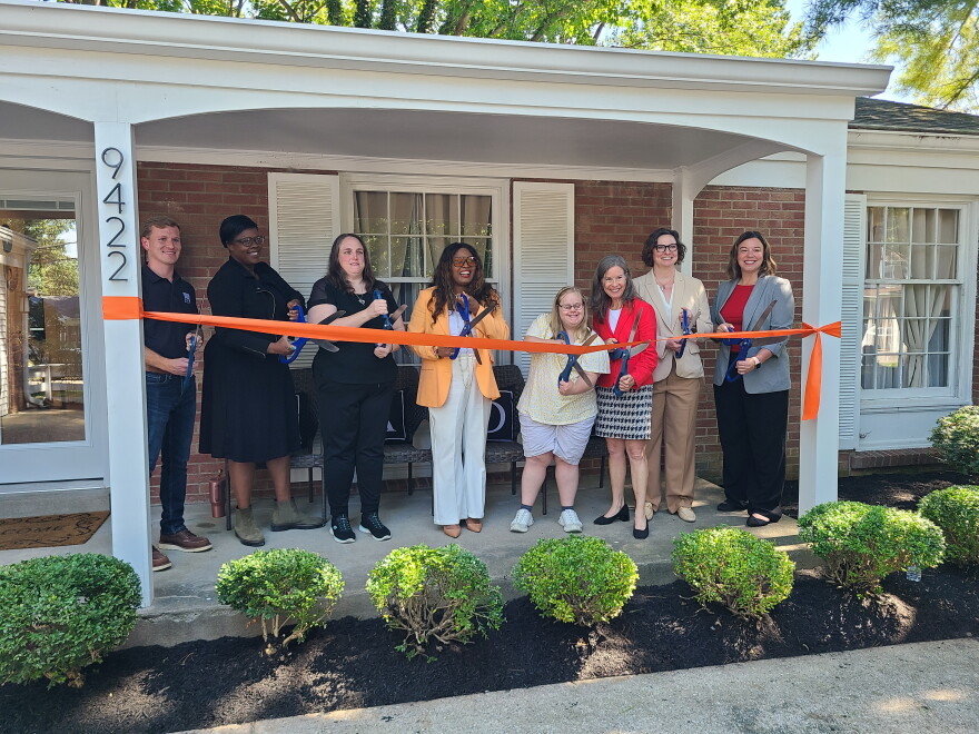 people stand on house porch and cut an orange ribbon with comically large scissors